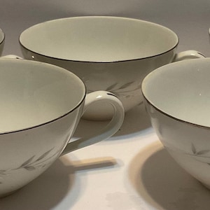 May include: Five white teacups with a delicate floral design and a thin silver rim. Each cup has a handle and sits on a white surface. The cups are arranged in a semi-circle, showcasing their elegant shape and design.
