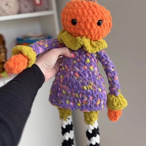 May include: A crocheted plush doll featuring an orange pumpkin head with a brown stem, a purple dress with yellow and orange speckles, a yellow ruffled collar, and long arms ending in orange cuffs. The legs are black and white striped with pink boots.