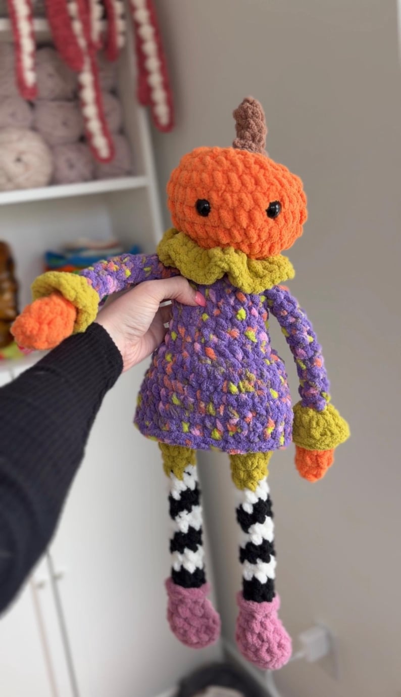 May include: A crocheted plush doll featuring an orange pumpkin head with a brown stem, a purple dress with yellow and orange speckles, a yellow ruffled collar, and long arms ending in orange cuffs. The legs are black and white striped with pink boots.