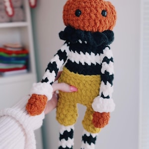 May include: A crocheted plush doll featuring a pumpkin-shaped head with a brown stem, black safety eyes, and a black ruffled collar. The body has black and white striped arms, mustard yellow shorts, and legs with black and white stripes and brown feet.