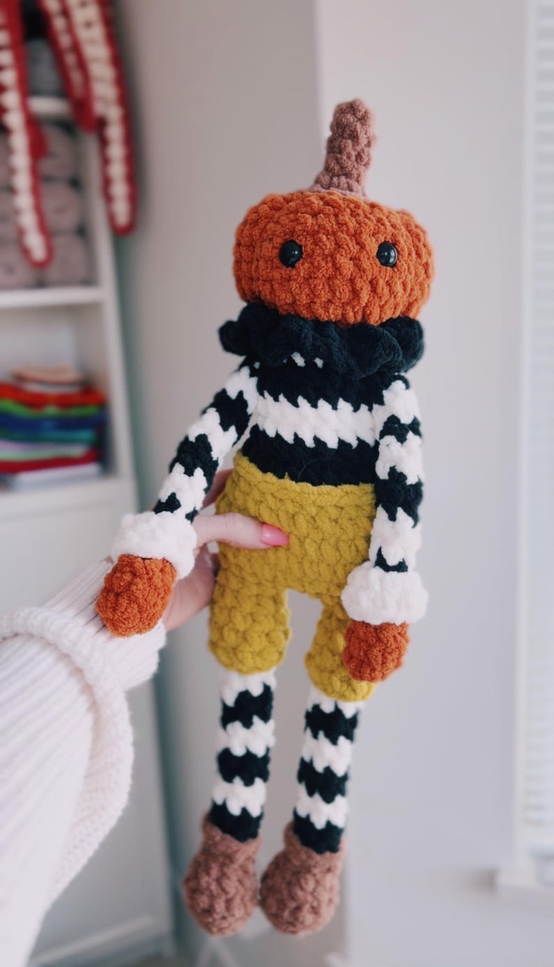 May include: A crocheted plush doll featuring a pumpkin-shaped head with a brown stem, black safety eyes, and a black ruffled collar. The body has black and white striped arms, mustard yellow shorts, and legs with black and white stripes and brown feet.