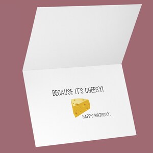 May include: A white greeting card with the text "BECAUSE IT'S CHEESY!" and "HAPPY BIRTHDAY." A yellow cheese wedge illustration is also featured on the card. The card is open, revealing the inside.