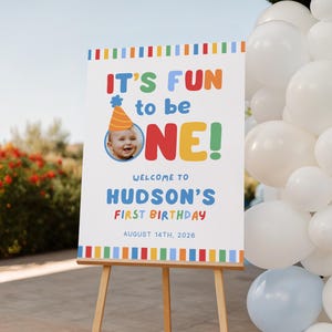 Pu&ograve; includere: Un cartello bianco su un cavalletto di legno con la scritta "IT'S FUN to be ONE!" in lettere colorate. Il cartello d&agrave; il benvenuto al primo compleanno di Hudson il 14 agosto 2026. &Egrave; inclusa una foto di un bambino con un cappello da festa.