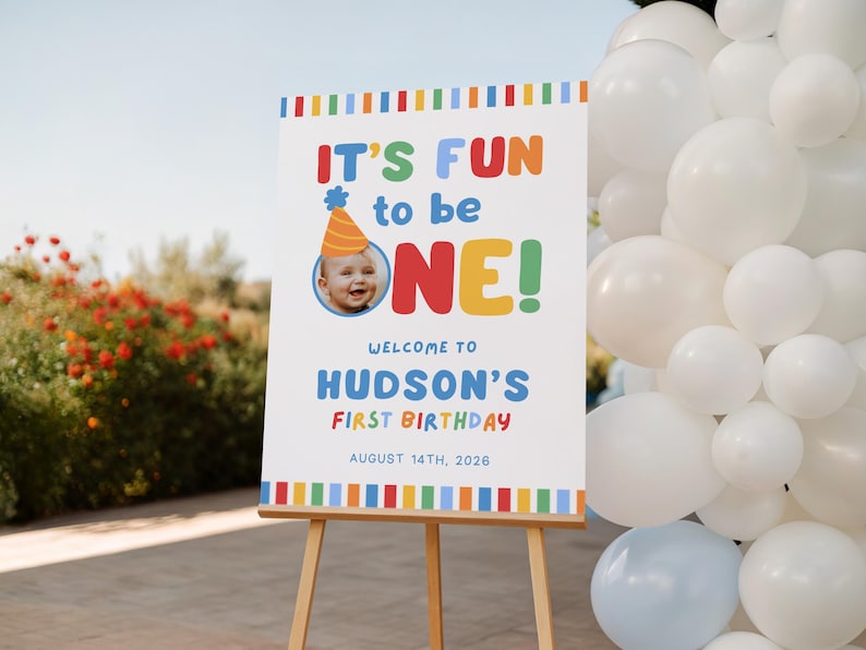 Pu&ograve; includere: Un cartello bianco su un cavalletto di legno con la scritta "IT'S FUN to be ONE!" in lettere colorate. Il cartello d&agrave; il benvenuto al primo compleanno di Hudson il 14 agosto 2026. &Egrave; inclusa una foto di un bambino con un cappello da festa.