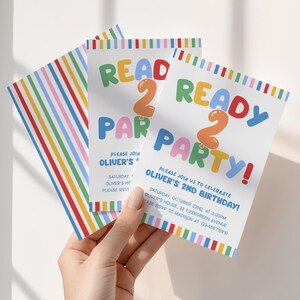 May include: Three white party invitations with colorful text and stripes. The invitations read "READ 2 READY PARTY!" and include party details for Oliver's 2nd birthday. The invitations are held by a hand.