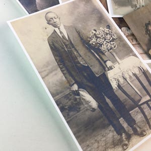 May include: A black and white photograph of a man in a suit holding a bouquet of roses. He is standing next to a small table with a lace tablecloth. The man is wearing a tie and a vest. The photo is in a vintage style.