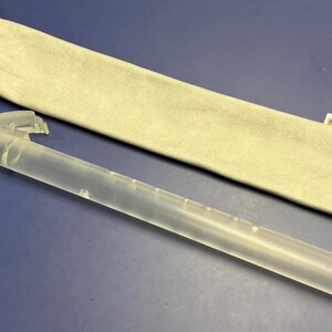 May include: A clear, cylindrical plastic instrument with measurement markings, next to a light beige fabric carrying case. The instrument has a small, clear plastic piece attached. The case has a small tag with text. The items are on a blue surface.