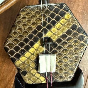 May include: A close-up of an erhu, a two-stringed bowed musical instrument. The hexagonal sound box has a snakeskin pattern in shades of brown and yellow. The bow is made of wood and horsehair. The instrument rests on a wooden surface.