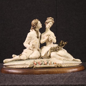 Pode incluir: Estatueta de porcelana retratando um casal sentado em trajes de época. O homem, com um casaco branco, olha para a mulher, que usa um vestido branco. A base é de madeira oval com detalhes florais.