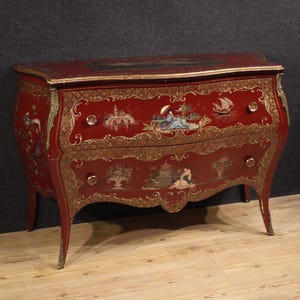 May include: A red antique chest of drawers with two curved drawers. The chest is adorned with gold trim and painted scenes of figures and landscapes. The legs are curved and the hardware is round.
