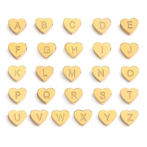 May include: A collection of twenty-six gold-colored heart-shaped charms, each featuring a single embossed letter of the alphabet from A to Z. These small, uniform metal pieces are arranged in a grid pattern, displaying a smooth, polished finish.