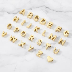 May include: A collection of gold-colored metal alphabet letter beads from A to Z, each featuring a small hole for threading. The set includes twenty-six individual letter charms and one small gold-colored heart-shaped charm arranged in rows.