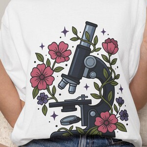 Op de afbeelding: Een wit t-shirt met korte mouwen en een grafische print van een donkerblauwe microscoop verweven met roze en paarse bloemen, groene bladeren en kleine paarse steraccenten. Het ontwerp is gecentreerd op de borst van het kledingstuk.