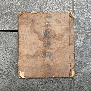 May include: A worn, brown, paper-covered book with the Japanese text "日本 陸軍" (Japan Army) printed on the cover.