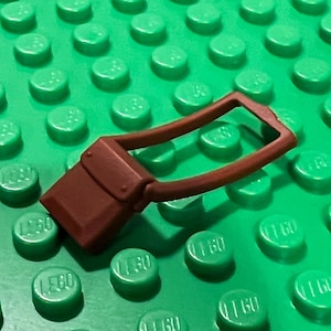 May include: A brown Lego purse with a handle, set against a green Lego baseplate. The purse is a reddish-brown color and has a rectangular shape with a curved handle.