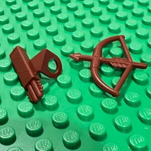 May include: A brown toy bow and arrow set with a quiver, resting on a green Lego baseplate. The bow has a detailed design, and the quiver holds several arrows. The set is designed for small figures.