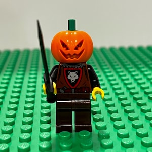 May include: A Lego minifigure with a jack-o'-lantern head, black torso, and brown vest with a wolf emblem. The figure holds a black sword and has yellow hands. It stands on a green Lego baseplate.