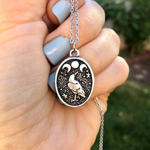 May include: A silver chain necklace with a silver oval pendant featuring a stylized raven with three crescent moons and stars on a black background.