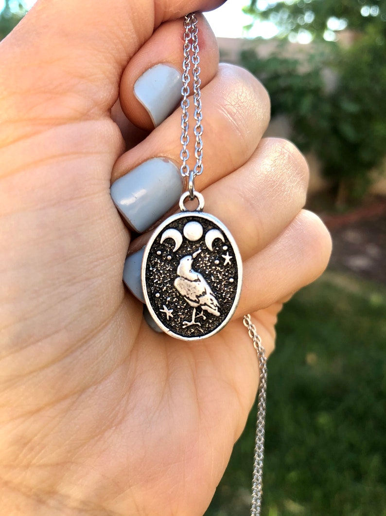 May include: A silver chain necklace with a silver oval pendant featuring a stylized raven with three crescent moons and stars on a black background.