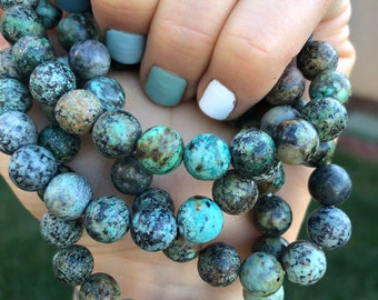 May include: A close-up of a turquoise and brown beaded bracelet. The bracelet is made of round beads with a speckled pattern. The bracelet is being held by a hand with light blue and white nail polish.