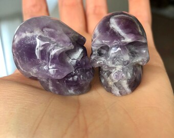 May include: Two amethyst crystal skulls, one with a more pronounced swirl pattern, are displayed on a hand. The skulls are carved from purple amethyst with white streaks.