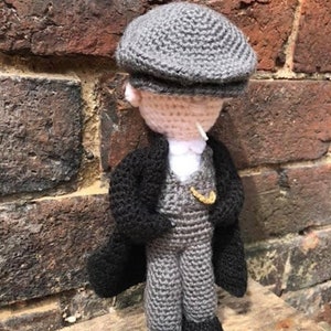 Op de afbeelding: Een gehaakte pop met een grijze pet, een zwarte jas en een grijze broek. De pop staat op een houten oppervlak.