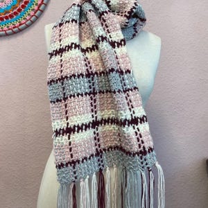Può includere: Sciarpa fatta a mano con motivo a quadri, esposta su un manichino. La sciarpa presenta una palette di colori rosa chiaro, grigio, bianco e bordeaux. La sciarpa ha frange alle estremità.