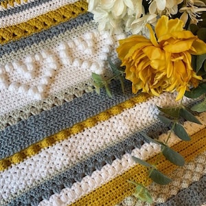 Puede incluir: Una manta de ganchillo con rayas de hilo blanco, gris y amarillo. La manta tiene un patrón texturizado con puntos y abalorios en relieve. La manta está sobre una superficie de madera con flores encima.