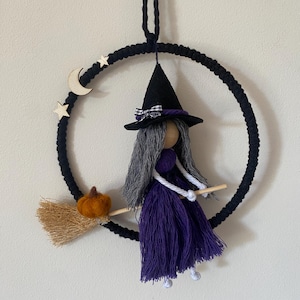May include: A hanging wall decoration featuring a black macrame hoop adorned with wooden star and moon cutouts. A doll with grey yarn hair, a black felt hat with a plaid bow, and a purple yarn dress sits on a wooden broomstick with a felt pumpkin.