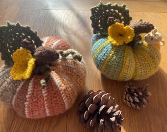 May include: Two handmade crochet pumpkins in shades of orange, brown, yellow, and green sit on a wooden surface. Each pumpkin has a crocheted leaf and flower detail. Two pine cones are also visible.