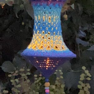 Peut inclure: Une lanterne crochetée à la main, dans des tons de bleu, turquoise et violet, suspendue à un arbre. La lanterne a une forme de cloche avec un pompon décoratif et des perles. La lanterne est éclairée de l'intérieur, créant une lueur chaleureuse.