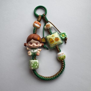 May include: A decorative keychain featuring a brown-haired doll figure in green clothing, with a green and brown braided cord. The keychain includes various charms, such as a cat, a cloud, a square with pear graphics, and a bead with the text "lucky day".