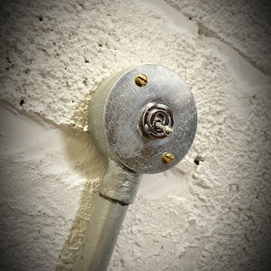 May include: A silver light switch with a round, metal cover and a brass toggle. The switch is mounted on a white brick wall with a silver metal pipe extending from the bottom.