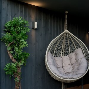 May include: A grey hanging egg chair with a grey cushion, hanging from a chain in front of a dark grey wooden wall. A small potted plant is on the left side of the image.