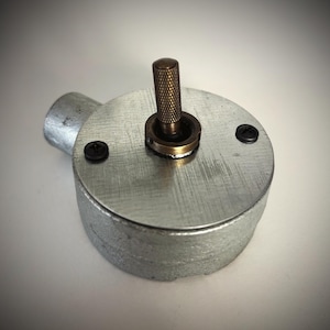 May include: A circular silver-coloured metal electrical switch box featuring a textured finish. The device includes a central vertical brass-toned knurled toggle switch and two small black screws securing the top plate to the main housing.