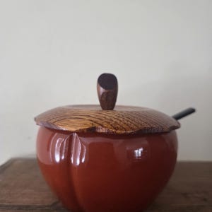 以下が含まれることがあります： 木製の蓋と小さな木製のハンドルが付いた、赤いリンゴ型の容器。 蓋には天然の木目模様があります。 小さな黒いスプーンが横にあります。 容器は木製の表面に置かれています。