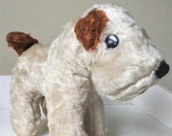 Può includere: Un cane di peluche vintage con pelliccia bianca, orecchie marroni e occhi neri. Il cane è in piedi su una superficie bianca.