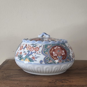 May include: A round, lidded ceramic container with a floral design. The container has a white base with a scalloped edge, and the lid has a blue rim and a small handle. The floral pattern includes red, orange, and green accents.