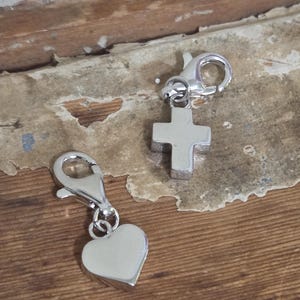 May include: Two silver charms with lobster clasps. One charm is a heart shape, and the other is a cross. The charms are displayed on a wooden surface with a vintage paper background. The charms are likely for a bracelet or necklace.