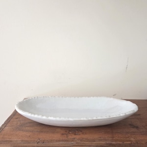 May include: An oval-shaped white ceramic dish with a textured, beaded rim. The dish has a smooth, glossy interior surface and a shallow basin, presenting a clean, neutral aesthetic suitable for serving or display purposes in any home setting.