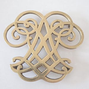 May include: A gold-colored, ornate metal object with a symmetrical design. The object features a central, stylized letter 'A' shape, surrounded by swirling, openwork patterns. The overall form is reminiscent of a decorative emblem or emblem.
