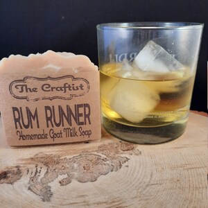 May include: A bar of goat milk soap with a brown paper label that reads "The Craftist Rum Runner Homemade Goat Milk Soap". The soap is sitting on a wooden surface next to a glass of amber liquid with ice cubes.