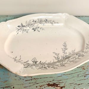 May include: An antique white oval serving platter with a floral design in gray. The platter has a scalloped edge and a slightly raised rim. The floral pattern is delicate and covers the edges of the platter.