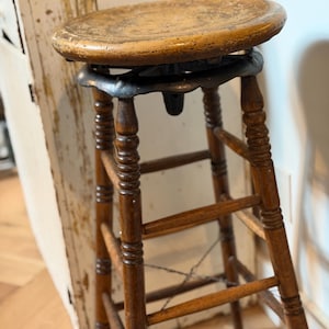 May include: A tall wooden stool featuring a round, worn light-brown seat and a dark-brown base with four turned legs. The seat is connected to the legs by a black metal swivel mechanism. Horizontal rungs connect the legs for stability.