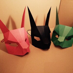May include: Three paper masks, one red, one black, and one green. The masks are all animal-like with pointy ears and are made of folded paper.