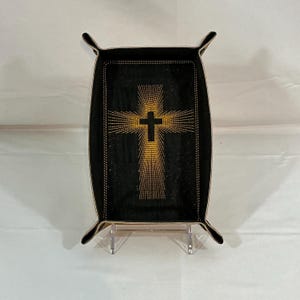 May include: Black leather tray with a gold embroidered cross design. The cross is centered on the tray and has a radiant pattern emanating from it. The tray has a rectangular shape with curved edges and a stitched border.