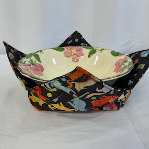May include: A floral patterned bowl rests inside a black and colorful fabric bowl cozy. The cozy has a geometric design with pointed corners. The bowl has a cream background with pink flowers and green leaves.