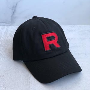 Könnte beinhalten: Schwarze Baseballkappe mit einem roten, gestickten "R" auf der Vorderseite. Die Kappe hat einen gebogenen Schirm und einen Knopf oben. Der Hut ist aus einem weichen Material.