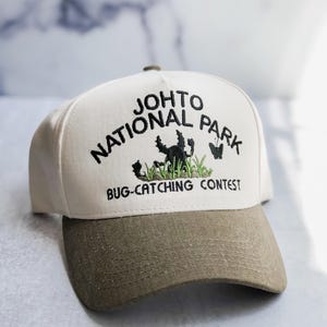 May include: A cream-colored baseball cap with an olive green brim. The front features black embroidered text reading Johto National Park Bug-Catching Contest, with a green embroidered patch of grass, a black beetle, and a small butterfly.