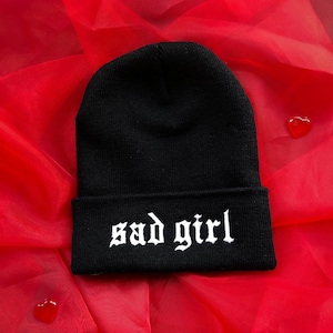 May include: Black beanie with white embroidered text "sad girl" on a red background.
